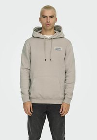 Felpa leggera beige con tasca frontale, cordini e logo sul petto. Realizzata in materiale morbido, presenta una vestibilità rilassata.