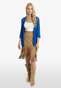 Zila mežģīņu kardigāns virs balta krekliņa, kopā ar brūnas zamšādas svārku ar frizūru un brūniem kovboju zābakiem, kam ir sarežģīta izšuvuma detaļa.