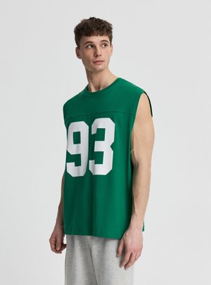 Giovane uomo con una maglietta verde senza maniche con il numero bianco 93, in piedi e che guarda di lato su uno sfondo semplice.