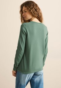 Helles grünes Sweater mit langen Ärmeln und rundem Ausschnitt, ausgestattet mit einer glatten Strickstruktur. Kombiniert mit hellblauen Jeans.