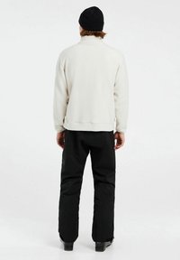 Hvid fleece-pullover med høj krave, parret med sorte bukser. Har en afslappet pasform og tekstureret overflade. Bæres med en sort hue.