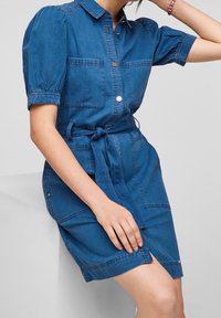Robe en denim bleu foncé avec de courtes manches bouffantes, un devant à boutons, deux poches poitrine et une ceinture nouée à la taille. Texture lisse, design décontracté.