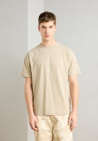 T-shirt en coton beige avec un col rond et des manches courtes, arborant un imprimé graphique vert clair centré sur la poitrine. Assorti à un short camo beige.
