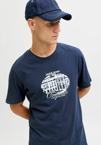 Camiseta de algodón azul marino con un estampado gráfico blanco de un edificio y texto. Combinada con una gorra de mezclilla oscura. Manga corta, corte relajado.