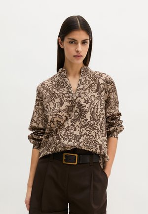Vrouw draagt een beige blouse met bruine bloemenpatronen, ingestopt in hooggetailleerde donkerbruine broek met een zwarte riem en gouden gesp.