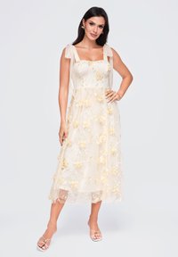 Femme en robe blanche sans manches avec broderie florale, debout la main sur la hanche devant un fond uni.