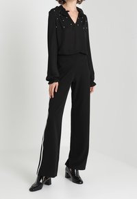 Musta paita, jossa on röyhelökaulus ja helmikoristelu, yhdistettynä mustiin leveälahkeisiin housuihin, joissa on valkoiset sivuviivat. Kiiltävät mustat korkokengät.