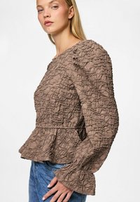 Blusa marrone strutturata con maniche a sbuffo e orlo peplum. Il tessuto presenta un design stropicciato e ha una vestibilità ampia. Abbinata a un denim.