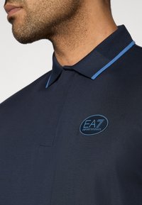 Μπλε πόλο μπλουζάκι με υφασμένη γιακά και ανοιχτό μπλε περίγραμμα. Διαθέτει κυκλικό μπλε λογότυπο με την ένδειξη "EA7 Emporio Armani" στο στήθος.