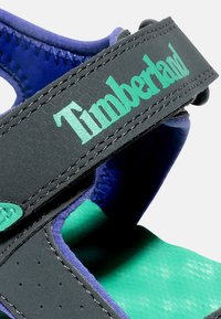 Musta sandaali turkoosi- ja sinivihreillä yksityiskohdilla, jossa on pehmustettu hihna, jossa lukee "Timberland" vihreällä, teksturoitu pohja ja sileä pinta.