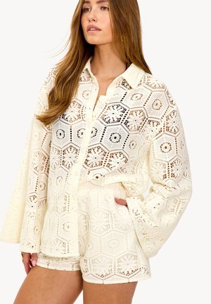 Vrouw draagt een crèmekleurig, met haakpatroon gebreid, lang mouwig overhemd met knopen en bijpassende shorts, met één hand in haar zak en kijkt iets naar de zijkant.