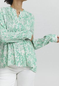Green floral-patterned blouse with a buttoned, round neckline, long sleeves, and pleated detailing. Made of lightweight fabric.