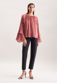 Blusa rosa con scollatura e maniche svasate, abbinata a jeans neri a gamba dritta e scarpe con tacco nere. Tessuto morbido.