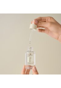 Heldere glazen fles met een wit en beige dop, gevuld met lichte olie. Een pipet wordt gebruikt om olie te doseren. Labels in zilveren en gouden lettertypes.