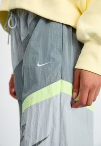 En persons hand vilar bredvid grå Nike träningsbyxor med diagonala neongr öna ränder och en gul sweatshirtärm synlig.