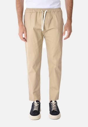 Uomo che indossa pantaloni beige con coulisse, t-shirt bianca e scarpe sportive nere con lacci bianchi, in piedi davanti a uno sfondo semplice.