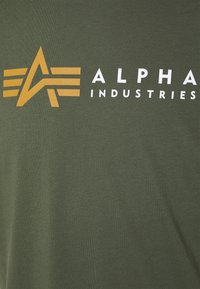 Olivengrønn T-skjorte med korte ermer som har en fremtredende gul og hvit Alpha Industries-logo med et stilisert pil-design.