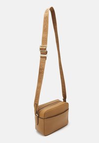 Kožená ramenní taška v tan barvě s obdélníkovým tvarem, zipovým uzavíráním a logem vyraženým na popruhu. Hladká textura s jemnými švy.