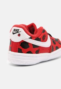 Červené kožené tenisky s černými skvrnami, bílými akcenty a texturovanou podrážkou. Obsahují logo Nike a malý detail berušky na patě.