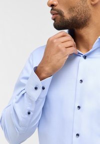 Hellblaues, strukturiertes Hemd mit taillierter Passform, Button-Down-Kragen und marineblauen Knöpfen. Die Manschetten sind geknöpft und zeigen einen maßgeschneiderten Look.