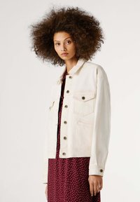 Balta džinsinė striukė su sagų užsegimu, dvi priekinės kišenės ir laisvas siluetas, dėvima ant bordo spalvos suknelės su baltais taškeliais.