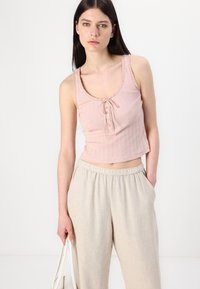 Top scurt din bumbac roz deschis, cu un model ribatat și margini ondulate, cu un detaliu de legare în față și nasturi, asortat cu pantaloni bej cu talie înaltă.