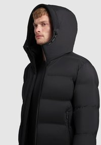 Black puffer jacket with a high collar and large hood, featuring quilted patterns, a front zipper, and side pockets. Smooth texture.