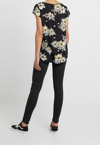 Blusa negra con flores amarillas y blancas, de mangas cortas y un detalle de ojal en la espalda, combinada con jeans ajustados negros y zapatillas.