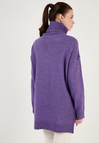Vijolični pulover s turtleneck ovratnikom iz pletenine, z udobnim krojem, z ribanim robom in daljšim hrbtom z stranskimi razpoki, prikazan z belimi hlačami.