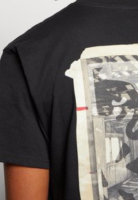 Svart bomulls-t-shirt med ett stort grafiskt tryck på baksidan, som visar en texturerad, monokrom design med röda accenter.