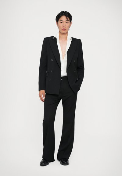 Sacou negru cu două rânduri de nasturi și rever ascuțite, asortat cu o cămașă albă cu guler deschis și pantaloni negri evazați.