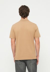 Άνδρας με κοντά σγουρά μαλλιά, φορώντας μπεζ πόλο με κοντά μανίκια και ξεθωριασμένο μαύρο τζιν, στέκεται με την πλάτη προς τα εμάς, σε απλό φόντο.