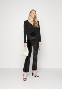 Top preto de mangas longas com detalhes franzidos à frente, combinado com calças pretas brilhantes e largas. Acessorizado com uma pequena bolsa creme.