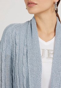 Cárdigan de punto azul claro con un patrón de cable, diseño de frente abierto y textura acanalada. Combinado con una blusa blanca de escote en V con letras plateadas.