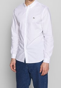 Camisa blanca de manga larga con botones, confeccionada en algodón. Presenta un cuello clásico y un pequeño logo bordado en verde en el pecho.