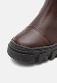 Bota de cuero marrón hasta el tobillo con punta redondeada, que presenta una suela de goma negra gruesa y detalles texturizados. Costura visible a lo largo del borde.