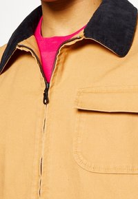 Tan canvas jacket with black corduroy collar, zip front, and chest pocket. Visible bright pink shirt underneath.