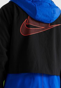 Svart jacka med blå huva som har en stor röd prickig Nike swoosh-logotyp på övre rygg.