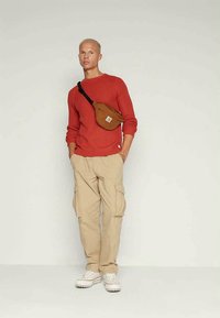 Rote gerippte Strickjacke, beige Cargo-Hosen mit seitlichen Taschen, braune Umhängetasche und weiße Sneakers mit strukturierten Sohlen.