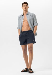 Mannequin portant une chemise rayée à boutons, un short de bain bleu foncé et des sandales noires. La chemise a les manches retroussées et une coupe décontractée.