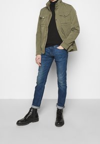 Olivgrön jacka med två bröstfickor, svart polotröja, blå jeans och svarta snörstövlar med grov sula.