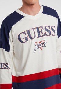 Weißes Langarmhemd mit blauen und roten Streifen, versehen mit einem strukturierten "GUESS"-Logo und einem V-Ausschnitt. Athletische Passform, weicher Stoff.