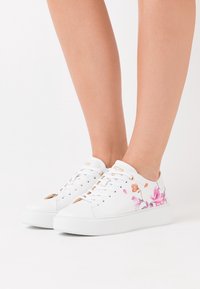 Baskets blanches avec motif floral, présentant des fleurs imprimées pastel en rose et orange, des accents dorés, et une semelle blanche texturée.