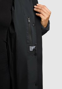 Čierna vetrovka s hladkým, matným povrchom. Má vrecko na zips a štítok s bielym textom na ľavej strane.