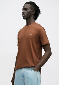 Brun bomulds-T-shirt med korte ærmer, rund halsudskæring og en lille broderet logo på brystet. Brugt med lyseblå denimbukser.