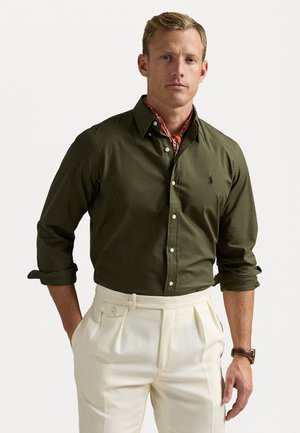 Camicia verde oliva con maniche arrotolate, abbinata a pantaloni plissettati di colore crema. Una sciarpa a fantasia è visibile attorno al collo.