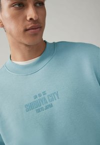 Svetlomodrá mikina s rebrovaným okrajom okolo krku. Na prednej strane je embosovaný nápis "SHIBUYA CITY TOKYO JAPAN". Mäkkučká, štruktúrovaná látka.
