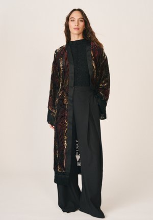 Femme debout portant un long manteau en velours à motifs, un haut noir texturé et un pantalon large noir sur fond uni clair.