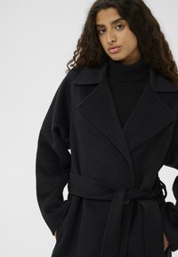 Palton negru din țesătură moale, având un guler larg, mâneci lungi și o curea asortată. Textură netedă cu un design minimalist.