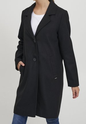 Femme portant un manteau noir boutonné jusqu'aux genoux, par-dessus une chemise blanche et un jean bleu, avec une main dans la poche du manteau.